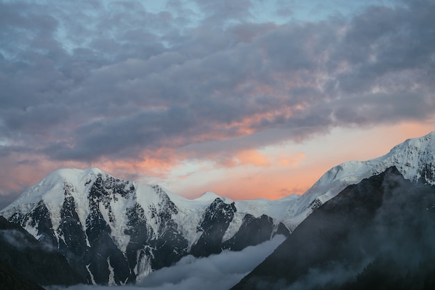 Paysage de montagne atmosphérique avec de grandes montagnes enneigées et des nuages bas dans la vallée sous un ciel nuageux à l'aube orange violet. Paysage alpin impressionnant avec un brouillard dense dans la vallée de montagne au coucher du soleil ou au lever du soleil