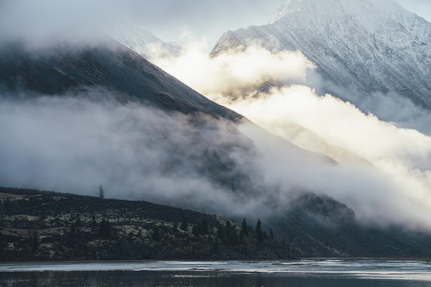 Paysage de montagne avec lac et silhouettes de conifères sur la colline moussue avec vue sur la haute montagne enneigée dans les nuages bas au soleil doré. Paysage en couches avec ombre et lumière du soleil dans les montagnes.