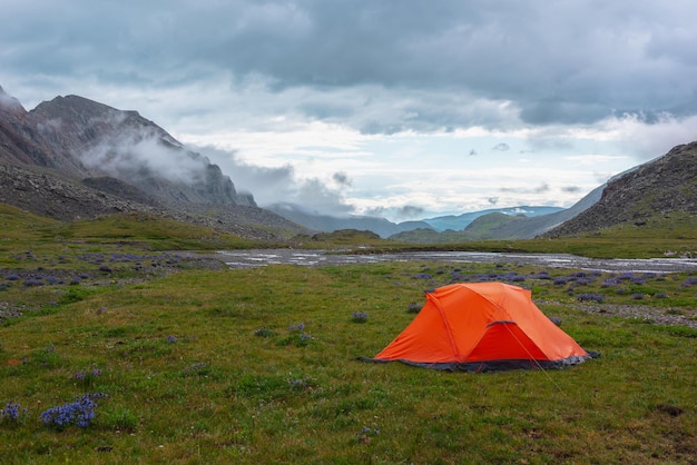 Photo paysage de montagne pittoresque avec une tente sous des gouttes de pluie au soleil sous un ciel nuageux paysage alpin spectaculaire avec une tente orange vif sur l'herbe parmi des fleurs violettes dans la vallée de la montagne verte par temps de pluie