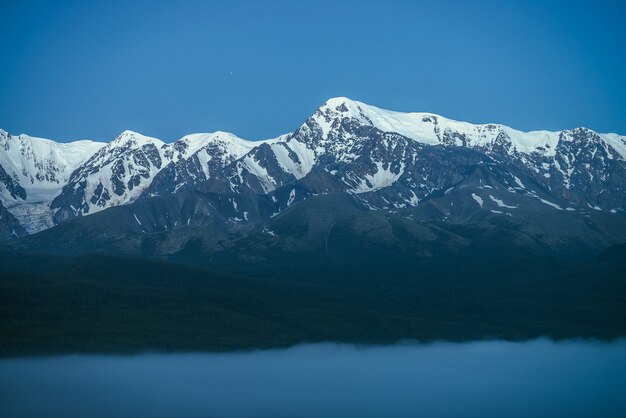 Paysage de montagnes atmosphériques avec un brouillard dense et une grande montagne enneigée sous un ciel crépusculaire. Paysage alpin avec de grandes montagnes enneigées sur un épais brouillard la nuit. Pinacle de neige élevée au-dessus des nuages au crépuscule.