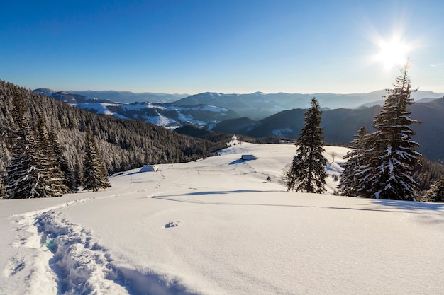 Paysage de Noël d'hiver de la vallée de la montagne par une journée ensoleillée et glaciale. Ancienne cabane de berger abandonnée en bois dans une neige blanche et propre, crête de montagne sombre et boisée, soleil éclatant sur fond d'espace de copie de ciel bleu.