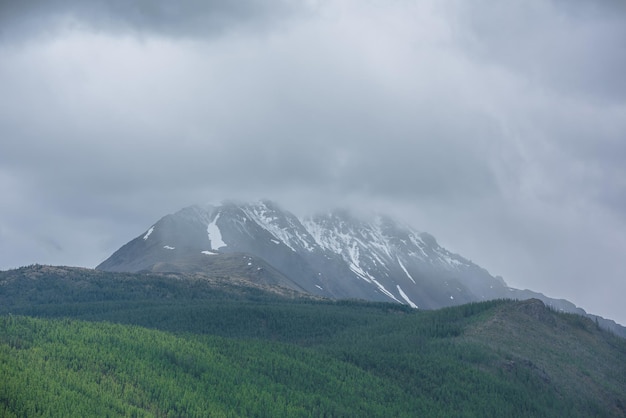 Paysage pluvieux atmosphérique simple avec une forêt verte ensoleillée à flanc de montagne et un sommet de montagne enneigé parmi des nuages bas gris Paysage sombre et couvert avec un sommet de montagne enneigé sous un ciel nuageux gris sombre