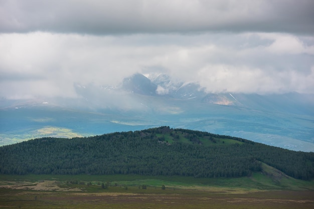 Paysage spectaculaire avec une colline de forêt verte avec vue sur la haute chaîne de montagnes enneigées ensoleillée dans des nuages bas pluvieux Paysage atmosphérique avec une forêt de conifères et de grandes montagnes enneigées par temps changeant