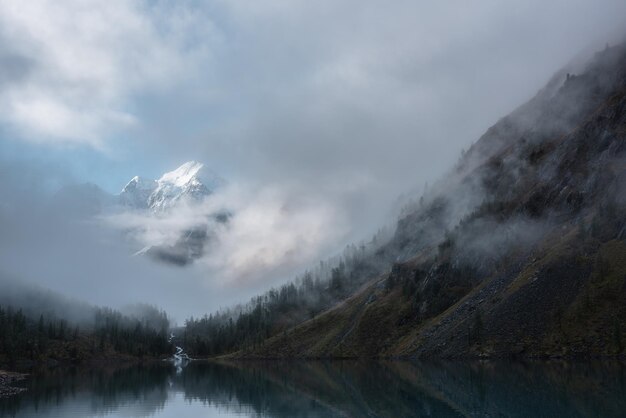 Paysage tranquille avec château de neige dans les nuages Un ruisseau de montagne coule des collines forestières dans un lac glaciaire Montagnes enneigées dans le brouillard Petite rivière et conifères se reflétant dans un lac alpin calme