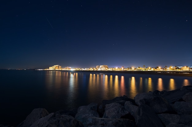 Paysage urbain de nuit avec mer et rochers au premier plan et ciel étoilé de nuit, paysage urbain de nuit d'une ville espagnole. Destination de voyage en Europe, vue carte avec lumières de la ville et front de mer la nuit