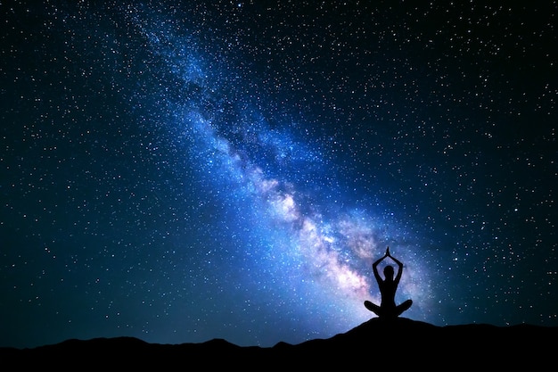 Paysage avec voie lactée bleue. Ciel nocturne avec des étoiles et la silhouette d'une jeune fille pratiquant le yoga dans la colline.