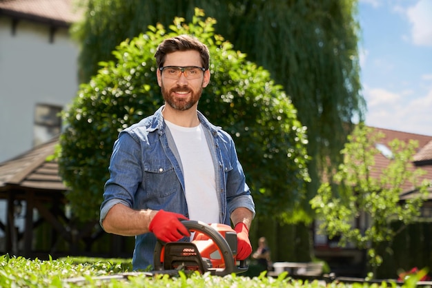 Paysagiste masculin souriant dans des lunettes de sécurité coupant le buisson avec taille-haie sur le portrait de l'arrière-cour