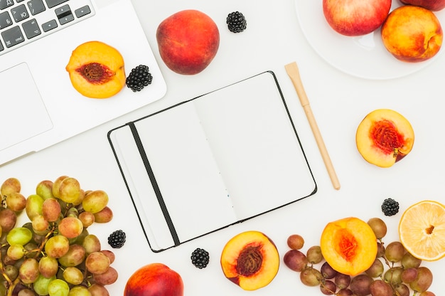 Pêche coupée en deux; raisins et mûres sur ordinateur portable; Journal et stylo sur fond blanc