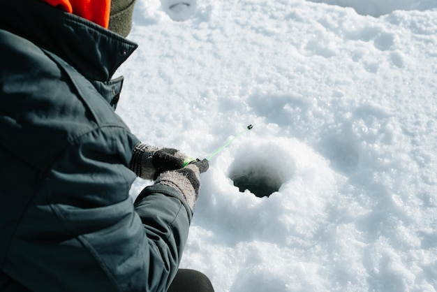 Pêche d'hiver, loisirs actifs, passe-temps et concept de sport. Gros plan sur un pêcheur assis sur de l'eau gelée près d'un trou percé dans la glace. Mise au point sélective sur les mains des hommes dans des gants tenant une canne à pêche