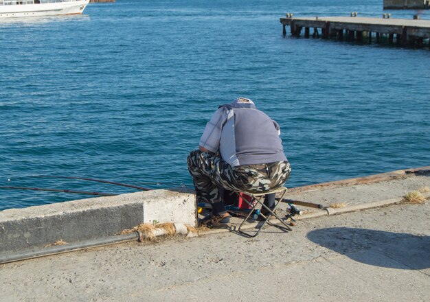 Un pêcheur âgé assis sur un parapet au bord de la mer et pêchant sur une canne à pêche