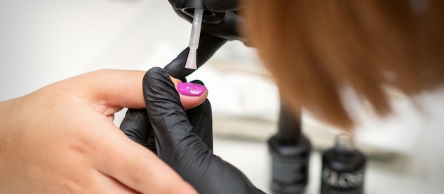 Peindre les ongles d'une femme. Mains de manucure en gants noirs appliquant du vernis à ongles rose sur les ongles des femmes dans un salon de beauté.