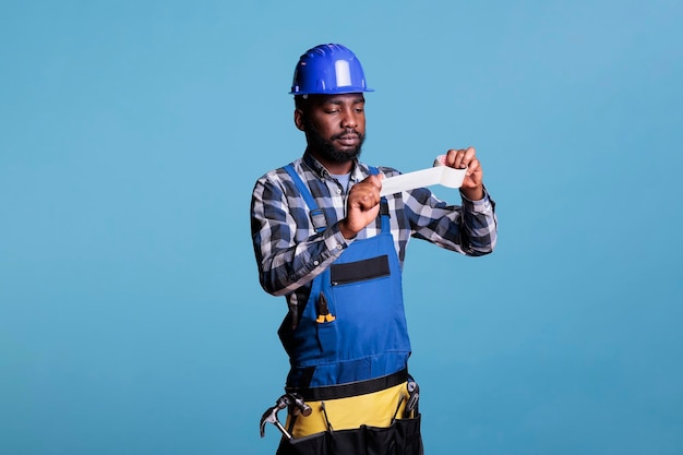 Le peintre en bâtiment professionnel tient un rouleau de papier peint à placer sur le mur avant de commencer à peindre. Travailleur de la construction portant une ceinture à outils de travail et un casque sur fond bleu.