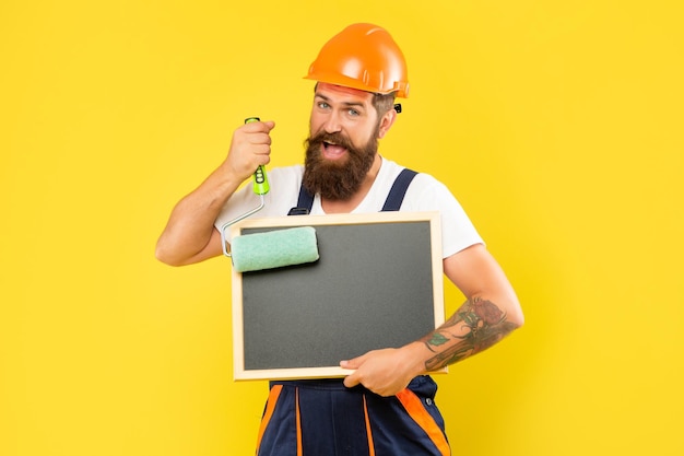 Un peintre en bâtiment souriant en casque et vêtements de travail tient un rouleau à peinture et un tableau noir