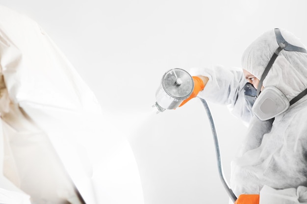 Peintre de carrosserie avec un pistolet à air comprimé pulvérisant de la peinture blanche sur une automobile dans la salle de peinture du service d'entretien de la voiture