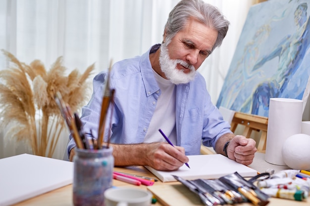 Le peintre dessine à la maison dans une pièce lumineuse, un homme barbu gris crée un chef-d'œuvre à l'aide d'un crayon