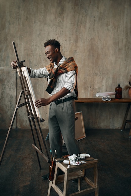 Un peintre heureux prépare un chevalet avec une toile dans un studio d'art. Un artiste masculin dessine sur son lieu de travail, un maître créatif travaille en atelier