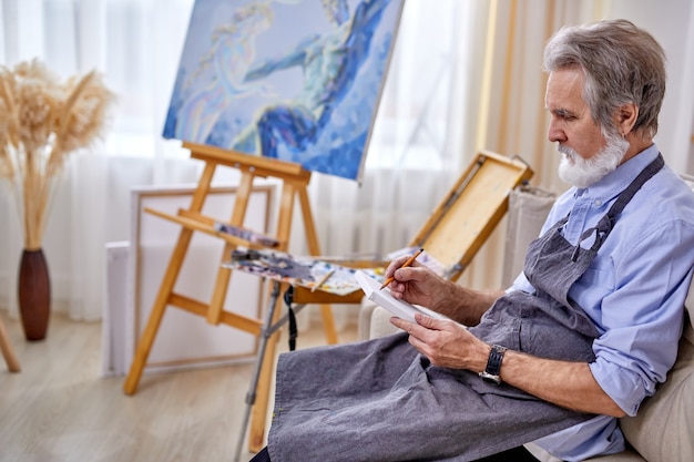 Peintre homme assis sur le canapé dessin au crayon, portant un tablier. dans un studio lumineux, lieu de travail