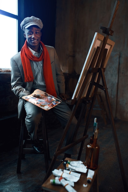 Photo peintre masculin à la mode avec des poses de palette au chevalet dans un studio d'art. l'artiste dessine sur son lieu de travail, le maître créatif travaille en atelier