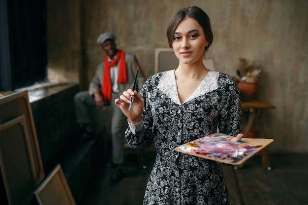 Peintre de portrait féminin détient palette et pinceau, modèle masculin en studio d'art. Artiste masculin debout sur son lieu de travail, maître créatif en atelier