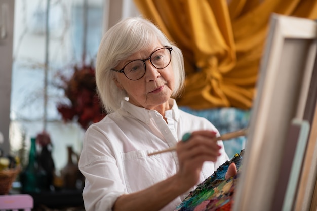 Peinture en atelier. Femme âgée aux yeux sombres, peinture en atelier se sentant bien et inspirée