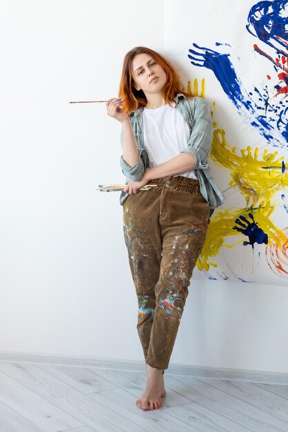 Peinture à la main Art-thérapie Loisirs relaxants Artiste féminine talentueuse et confiante avec des pinceaux debout devant des œuvres d'art abstraites colorées sur un mur blanc dans une galerie lumineuse