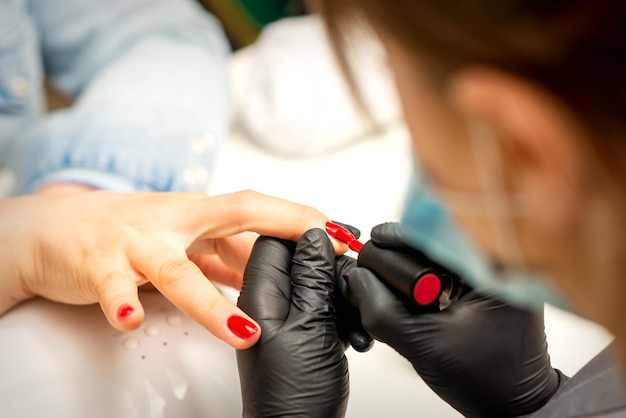Peinture de vernis de manucure Gros plan d'un maître de manucure portant des gants en caoutchouc noir appliquant du vernis rouge sur un ongle féminin dans le salon de beauté