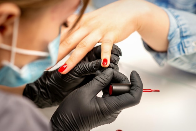 Peinture de vernis de manucure Gros plan d'un maître de manucure portant des gants noirs en caoutchouc et un masque de protection appliquant du vernis rouge sur un ongle féminin dans le salon de beauté