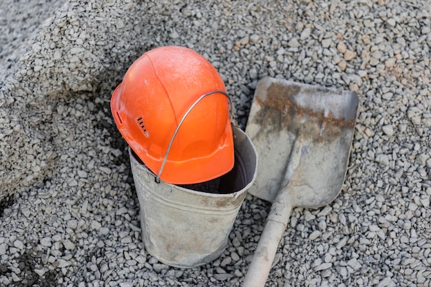 Une pelle enduite de béton dans un tas de gravats pour préparer le béton et un casque de construction orange d'un ouvrier se trouve sur un seau