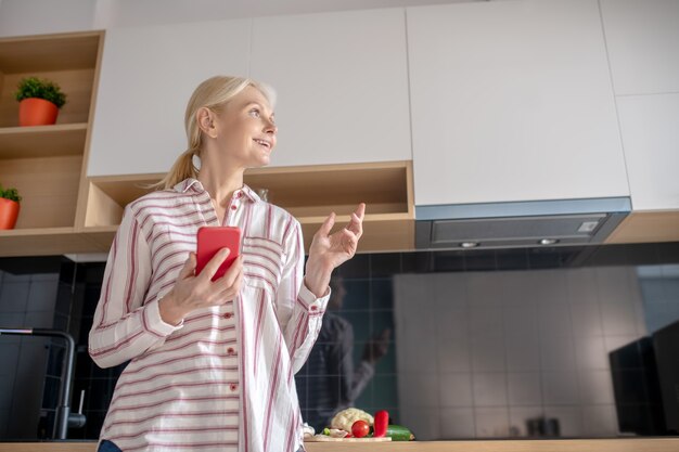En pensant. Femme debout dans la cuisine avec un smartphone à la main et à la réflexion