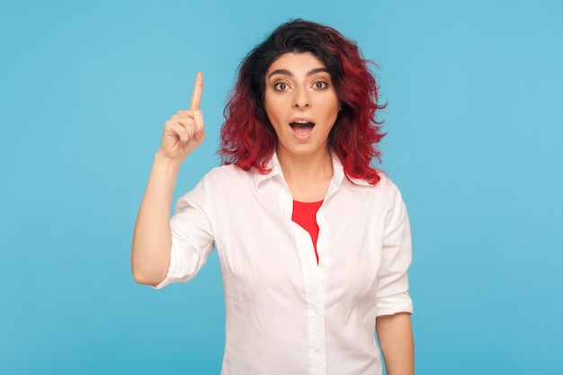 Pensée intelligente Portrait d'une fille hipster aux cheveux roux fantaisie en chemise pointant le doigt vers le haut avec une excellente idée inspirée par une solution de génie sachant répondre à la prise de vue en studio intérieur isolée sur fond bleu