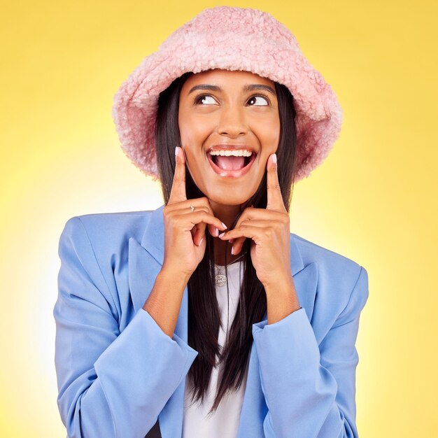 Photo pensée de mode et visage de femme sur fond jaune avec une expression drôle et ludique emoji bonheur et personne heureuse en studio sourire h dans un style de vêtements à la mode et une tenue avec un chapeau