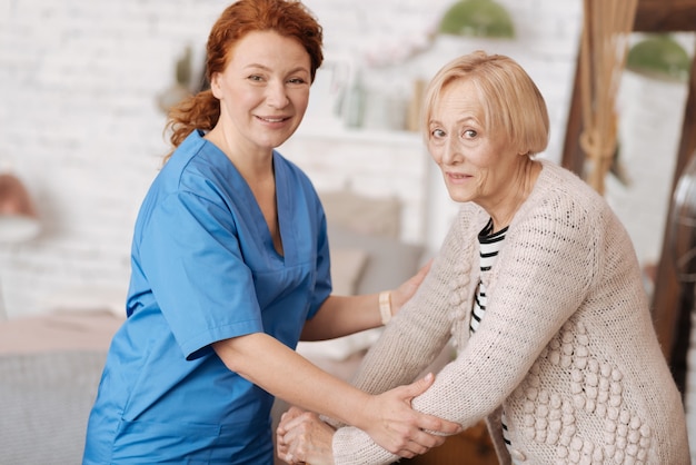 Penser aux seniors. Femme douce intelligente réussie tenant une dame âgée par ses mains pour s'assurer qu'elle se tient fermement tout en faisant une petite promenade autour de l'appartement