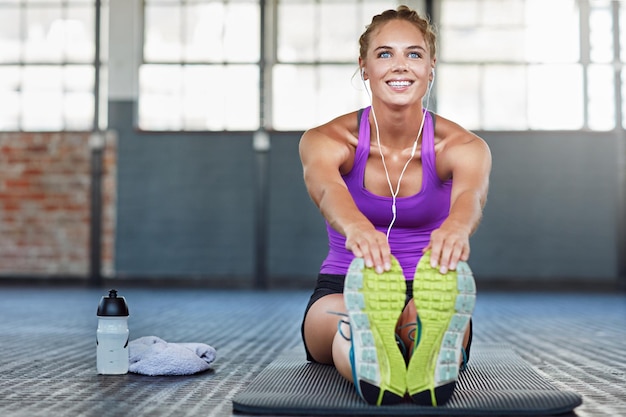 Penser à la forme physique et à la musique avec une femme qui s'étire dans une salle de sport tout en se préparant pour sa routine d'entraînement Exercice d'échauffement et écouteurs avec une jeune athlète féminine s'entraînant dans un centre sportif