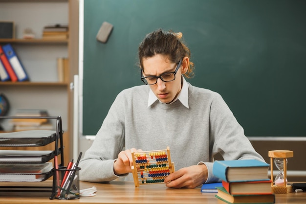 penser jeune enseignant portant des lunettes assis au bureau tenant et regardant l'abaque avec des outils scolaires en classe