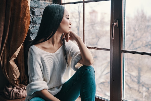 Penser à.... Jolie jeune femme gardant la main sur le menton et regardant ailleurs alors qu'elle était assise sur le rebord de la fenêtre à la maison