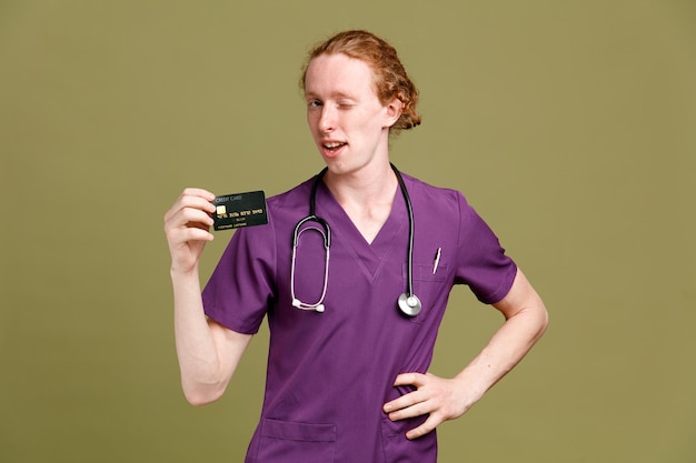 Penser mettre la main sur les hanches jeune médecin de sexe masculin portant l'uniforme avec stéthoscope tenant la carte isolé sur fond vert