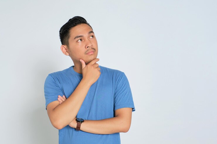 Pensif jeune homme asiatique en t-shirt bleu regardant l'espace de copie touchant son menton pensant et prenant une décision isolé sur fond blanc