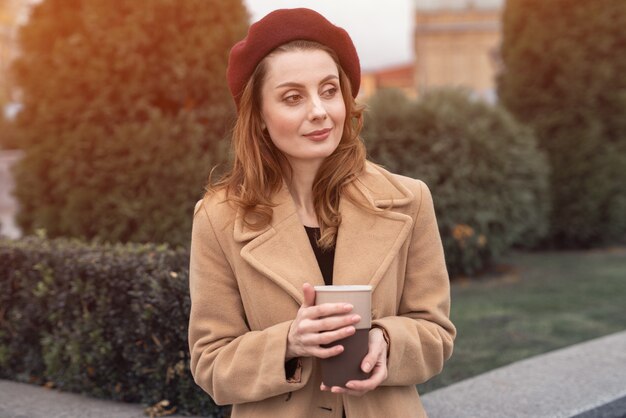 Pensive belle jeune femme tenant une tasse de café dans la rue. Concept de mode féminine