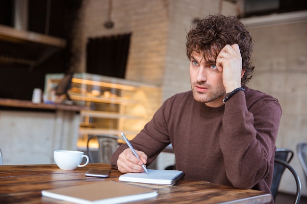 Pensive concentré beau jeune homme pensif bouclé en sweetshirt marron écrit dans un ordinateur portable assis dans un café et buvant du thé