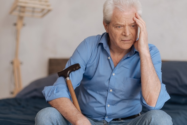 Pensive homme âgé sans joie tenant un bâton de marche et tenant sa tête tout en étant impliqué dans ses pensées