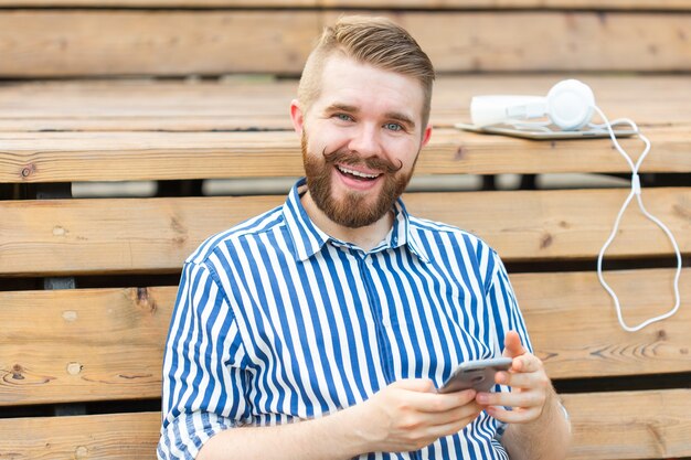 Pensive jeune étudiant hipster masculin sérieux avec une moustache et une barbe écrivant un message SMS à ses amis au repos après avoir étudié dans le parc, assis sur un banc en bois avec une tablette et des écouteurs.