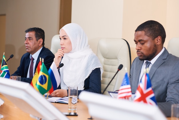 Pensive jeune femme déléguée en hijab à l'écoute de l'un de ses collègues rapport alors qu'il était assis à table entre deux hommes interculturels