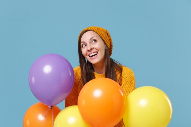 Pensive jeune femme fille en pull et chapeau posant isolé sur fond bleu. Fête d'anniversaire, concept d'émotions de personnes. Maquette de l'espace de copie. Célébrer tenir des ballons à air colorés en levant.