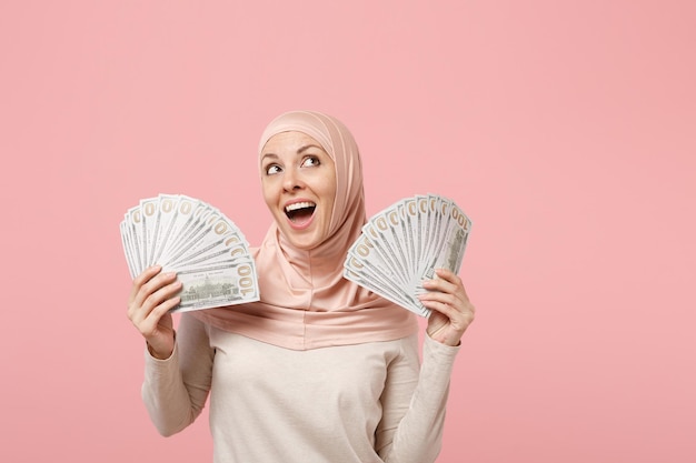 Pensive jeune femme musulmane arabe en vêtements légers hijab posant isolé sur fond rose. Concept de style de vie de l'islam religieux des gens. Maquette de l'espace de copie. Tenir un éventail d'argent liquide en billets de banque en dollars.