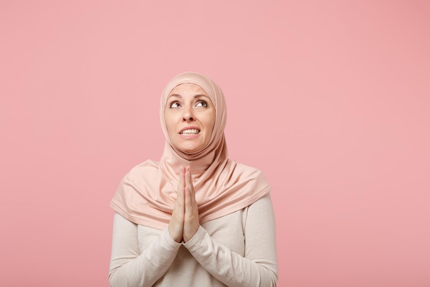 Pensive jeune femme musulmane arabe en vêtements légers hijab posant isolé sur fond rose en studio. Concept de style de vie de l'islam religieux des gens. Maquette de l'espace de copie. Tenir les mains jointes en prière.