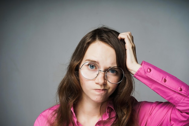 Pensive jeune fille aux yeux bleus dans une chemise rose et des lunettes regarde la caméra