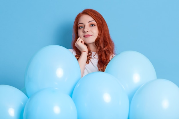 Pensive jeune fille européenne en chemise blanche isolée sur un mur bleu avec des ballons à air comprimé. Fête d'anniversaire. Jolie fille au gingembre garde le poing sous le menton et pense à la fête.