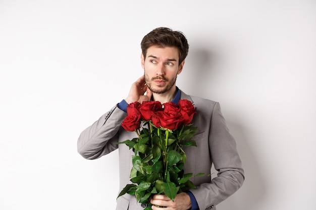 Pensive jeune homme en costume tenant un bouquet de fleurs, en attente de date le jour de la Saint-Valentin, debout sur fond blanc.