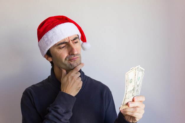 Photo pensive man with santa red hat holding dollar bills over white background personne regardant de l'argent