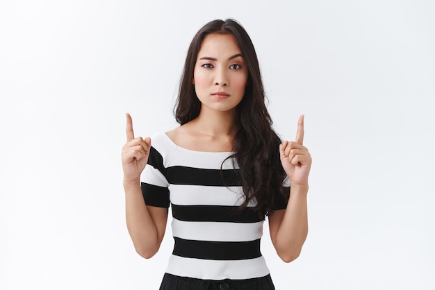 Pensive suspecte et sceptique jeune femme brune asiatique en t-shirt rayé levant un sourcil et regardant la caméra avec incrédulité levant les mains vers le haut pointant vers le haut promo debout fond blanc